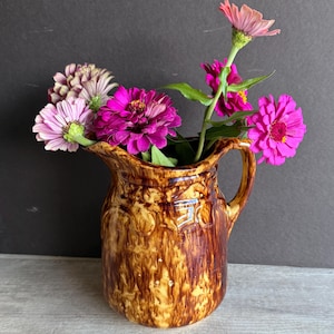 May include: A brown ceramic pitcher with a handle, filled with vibrant pink and purple zinnias. The pitcher has a textured, glazed finish with a decorative pattern. The flowers are in full bloom, adding a pop of color against the dark background.