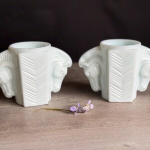 May include: Two white, Art Deco-style vases with horse head details. Each vase has a central column with a chevron pattern. A small sprig of purple flowers rests on the wooden surface between the vases.