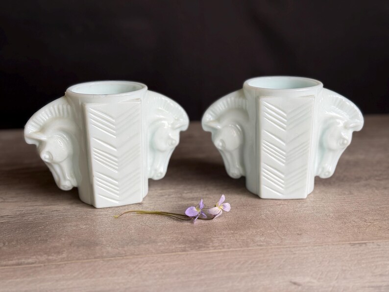 May include: Two white, Art Deco-style vases with horse head details. Each vase has a central column with a chevron pattern. A small sprig of purple flowers rests on the wooden surface between the vases.
