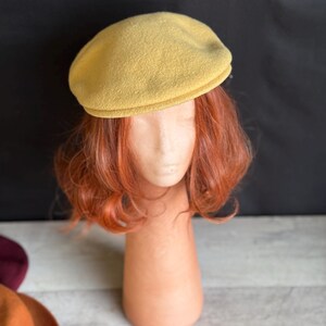 Puede incluir: Un sombrero tipo boina de color amarillo mostaza. El sombrero se exhibe en una cabeza de maniqu&iacute; con cabello rojo. El fondo es una mezcla de negro y blanco. Otros sombreros y hojas de oto&ntilde;o son visibles en el fondo.