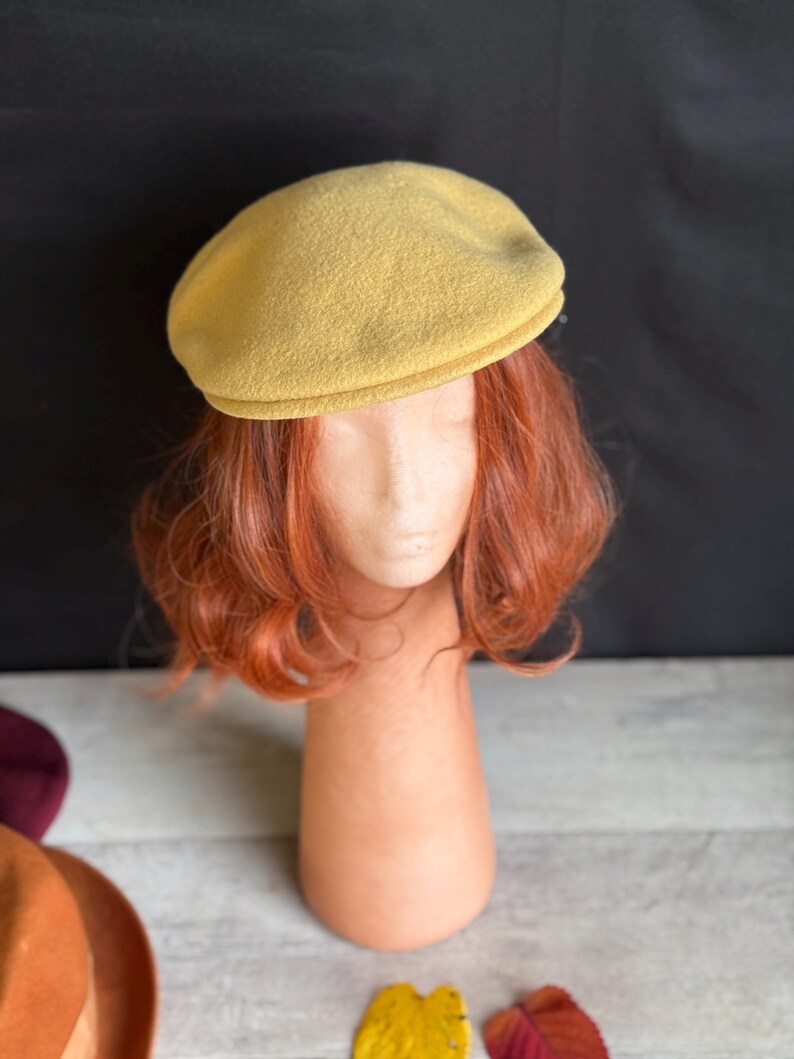 Puede incluir: Un sombrero tipo boina de color amarillo mostaza. El sombrero se exhibe en una cabeza de maniqu&iacute; con cabello rojo. El fondo es una mezcla de negro y blanco. Otros sombreros y hojas de oto&ntilde;o son visibles en el fondo.
