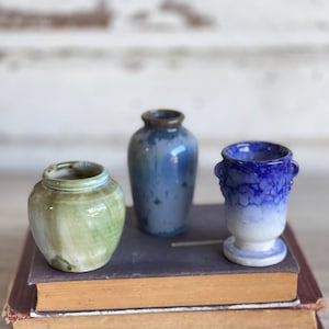 May include: Three small ceramic vases in different colors and styles. The vases are arranged on a stack of books. The vase on the left is green and white, the vase in the middle is blue and white, and the vase on the right is blue and white with a textured surface.