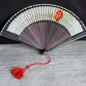 May include: A decorative folding fan with a red tassel. The fan features a floral design with a red flower and green leaves. The fan's ribs are a dark brown color, and the top edge has a white lace-like pattern.