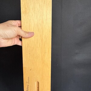 May include: A rectangular wooden object with a light brown finish, held by a hand. A green circular accent is near the top. The bottom has a carved-out section with three dots and the name "Roland Torner" is written on the side.