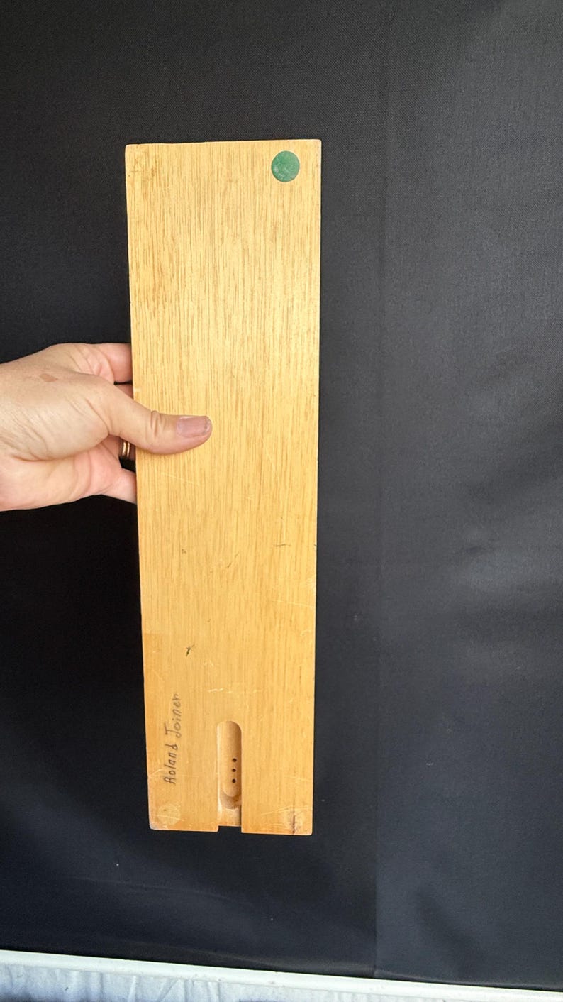 May include: A rectangular wooden object with a light brown finish, held by a hand. A green circular accent is near the top. The bottom has a carved-out section with three dots and the name "Roland Torner" is written on the side.