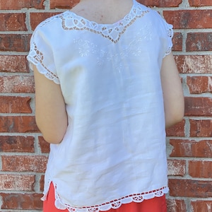 Puede incluir: Top blanco de lino con delicado ribete de encaje y bordado. El top tiene un escote redondo y mangas cortas.