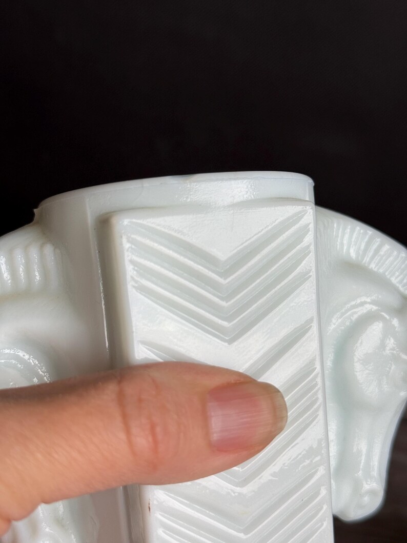 May include: A close-up of a white, Art Deco-style horse head figurine. The figurine features a geometric design with chevron patterns. The horse's head is visible, with details of the eye and mane. The figurine is made of a glossy, opaque material.