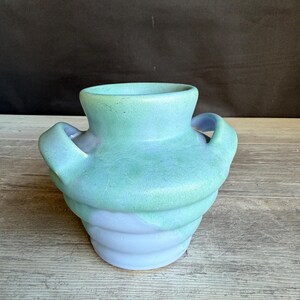 May include: A ceramic vase with a wide mouth and two side handles, featuring a ribbed body design. The upper portion is finished in a matte mint green glaze, while the lower section transitions into a smooth, pale lavender blue color.