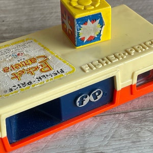 May include: A vintage yellow, orange, and blue Fisher-Price toy camera with a red lens and a yellow block with a starburst design on top. The camera has a blue and white label that reads "Fisher-Price Pocket Camera".