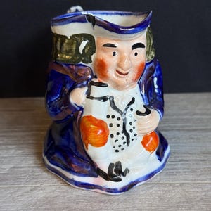May include: A ceramic pitcher in the shape of a man, featuring a blue coat, white shirt, and a tricorn hat. The pitcher has a handle and is painted with orange cheeks and a smiling face. The design is detailed and the colors are vibrant.