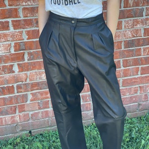 May include: Black leather pants with pleats and a button closure. The pants are a high-waisted style.