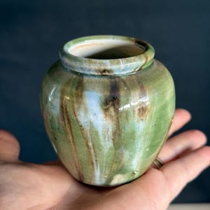 May include: A small, round ceramic vase featuring a glossy glaze in shades of sage green, light blue, and dark brown. The surface displays a subtle crackle texture with vertical drip patterns running down the sides toward the base.