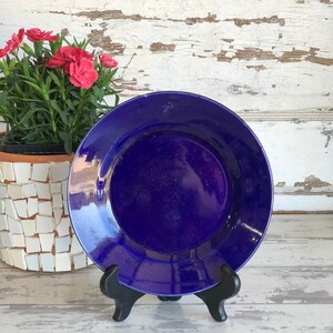 May include: A deep blue ceramic plate with a glossy finish, displayed on a black stand. The plate is round and has a simple design.