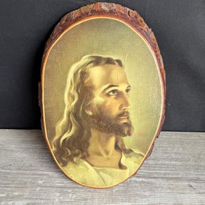 May include: Oval wooden plaque featuring a portrait of Jesus with long hair and a beard. The image is set against a golden background and framed by the natural edge of the wood, showcasing a rustic aesthetic.