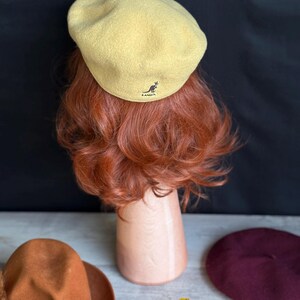 Puede incluir: Boina amarillo claro con el logo de Kangol, exhibida en una cabeza de maniqu&iacute; con cabello rojo. Tambi&eacute;n se ven un sombrero marr&oacute;n con una cinta y una boina burdeos. Hojas de oto&ntilde;o dispersas.