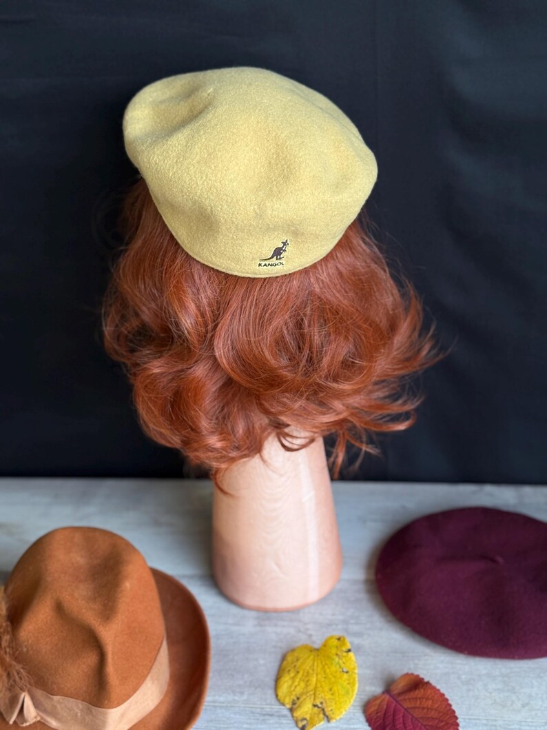 Puede incluir: Boina amarillo claro con el logo de Kangol, exhibida en una cabeza de maniqu&iacute; con cabello rojo. Tambi&eacute;n se ven un sombrero marr&oacute;n con una cinta y una boina burdeos. Hojas de oto&ntilde;o dispersas.