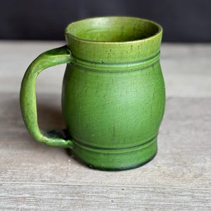 May include: A handcrafted, green ceramic mug with a curved handle. The mug has a textured surface and a slightly flared rim. The mug is sitting on a light-colored wooden surface.