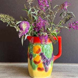 May include: A colorful ceramic pitcher with a red handle and rim, decorated with a fruit motif. The pitcher is filled with vibrant purple flowers and greenery, creating a striking contrast. The pitcher sits on a wooden surface against a dark background.