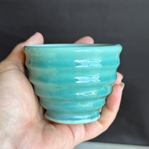 May include: A small, turquoise ceramic cup with a glossy finish and a series of horizontal, wavy ridges encircling the exterior. The cup features a flared rim and a narrow, circular base, showcasing a smooth, reflective surface texture throughout.