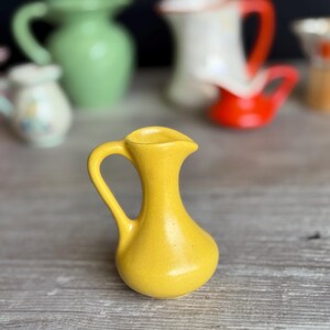 May include: A small, bright yellow ceramic pitcher with a smooth, matte finish. The vessel features a rounded base, a slender neck, and a curved handle on the side. The spout is slightly pointed, extending from the top rim of the pitcher.
