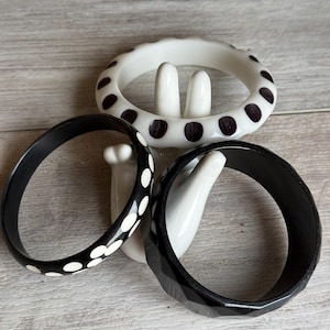 May include: Three bangle bracelets are displayed together. One is white with black polka dots, one is black with white polka dots, and the third is a solid black bangle with a faceted, geometric surface texture around the outer edge.