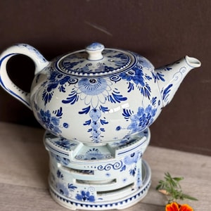 May include: A white and blue ceramic teapot with floral patterns, resting on a matching warmer. The teapot features a curved handle and spout. The warmer has a circular base with cutouts. A marigold flower and greenery are in the foreground.
