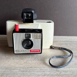 May include: Vintage Polaroid Swinger Model 20 camera. The camera has a black and silver face with a red accent and a black viewfinder. The camera is cream-colored and has a black wrist strap.