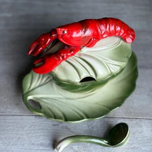 May include: A ceramic tureen shaped like a green cabbage leaf with a bright red lobster figurine attached to the lid. The set includes a matching green cabbage leaf saucer and a small green ladle with a curved handle, all featuring a glossy finish.