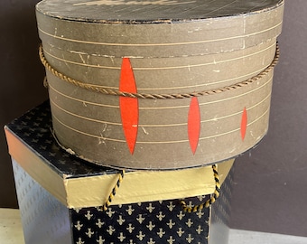 May include: Two vintage hat boxes stacked on top of each other. The top box is round and brown with red accents and a rope trim. The bottom box is black with a gold fleur-de-lis pattern.
