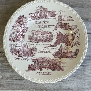 May include: A white ceramic plate with a brown design depicting various landmarks in Colorado Springs, including Seven Falls, Pikes Peak, the Broadmoor Hotel, and the Garden of the Gods. The plate has a rope-like border.