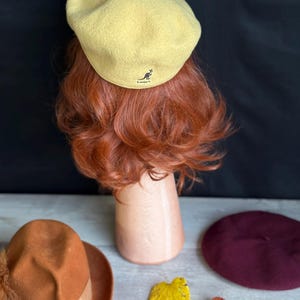 Puede incluir: Un boina amarillo claro con el logo de Kangol, exhibida en una cabeza de maniqu&iacute; con cabello rojo. Tambi&eacute;n se ven un sombrero naranja con pluma y una boina burdeos, junto con hojas de oto&ntilde;o.