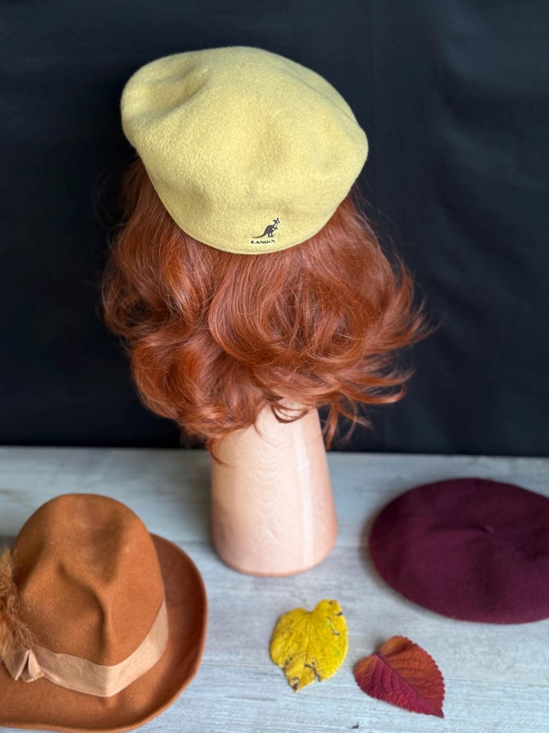 Puede incluir: Un boina amarillo claro con el logo de Kangol, exhibida en una cabeza de maniqu&iacute; con cabello rojo. Tambi&eacute;n se ven un sombrero naranja con pluma y una boina burdeos, junto con hojas de oto&ntilde;o.