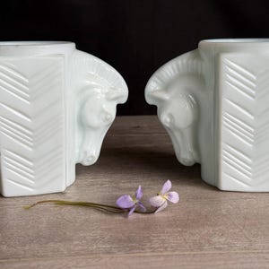 May include: Two white art deco horse head vases with a geometric design. The vases are placed on a wooden surface with a sprig of purple flowers.
