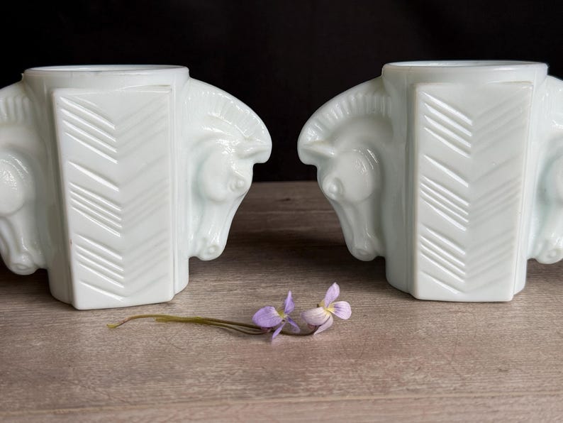 May include: Two white art deco horse head vases with a geometric design. The vases are placed on a wooden surface with a sprig of purple flowers.