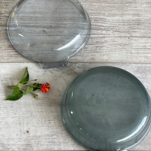May include: Two vintage, gray-tinted glass Pyrex lids and a matching dish. The lids are round with a handle on one side. The dish is also round and has a handle. A small sprig of flowers with red and yellow blooms is on the side.