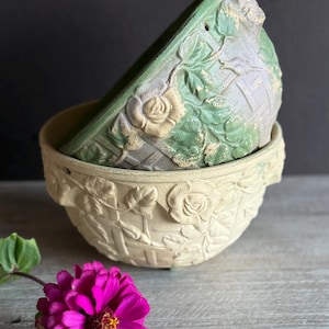 May include: Two decorative ceramic bowls, one nested inside the other. The bowls are cream and green with a floral and basket weave design. A bright pink flower with a yellow center rests on a wooden surface.