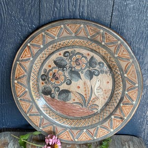 May include: A round ceramic plate featuring a hand-painted design of a bird perched near large flowers and foliage in shades of brown, gray, and cream. The wide rim is decorated with a repeating geometric pattern of triangles and white dots.