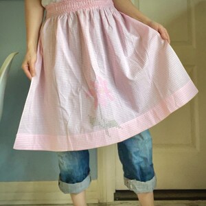 May include: A pink and white gingham apron with a floral embroidery design. The apron has a gathered waist and a pink border. The apron is a kitchen or craft accessory.