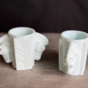 May include: Two white, vintage-style ceramic cups. One cup features a horse head design on each side, while the other has a single horse head handle. Both cups have a light blue interior and are set on a wooden surface.