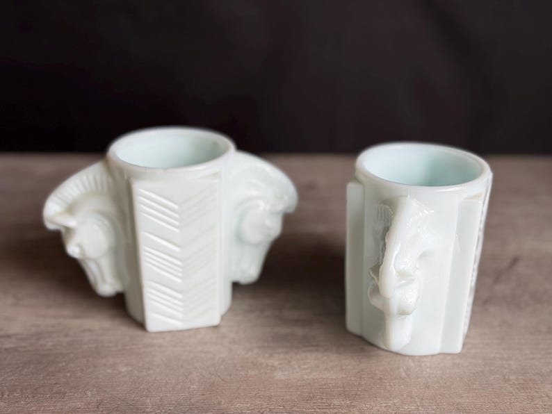 May include: Two white, vintage-style ceramic cups. One cup features a horse head design on each side, while the other has a single horse head handle. Both cups have a light blue interior and are set on a wooden surface.