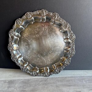 May include: A silver-colored, ornate serving tray with a detailed floral and scrollwork pattern. The tray has a scalloped edge with a raised, decorative border. The center of the tray is smooth and reflective, showing signs of wear.