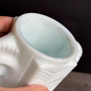 May include: A white, vintage-style creamer with a geometric design. The creamer has a square base and a rounded top, with a small handle on the side. The interior is a pale blue color.