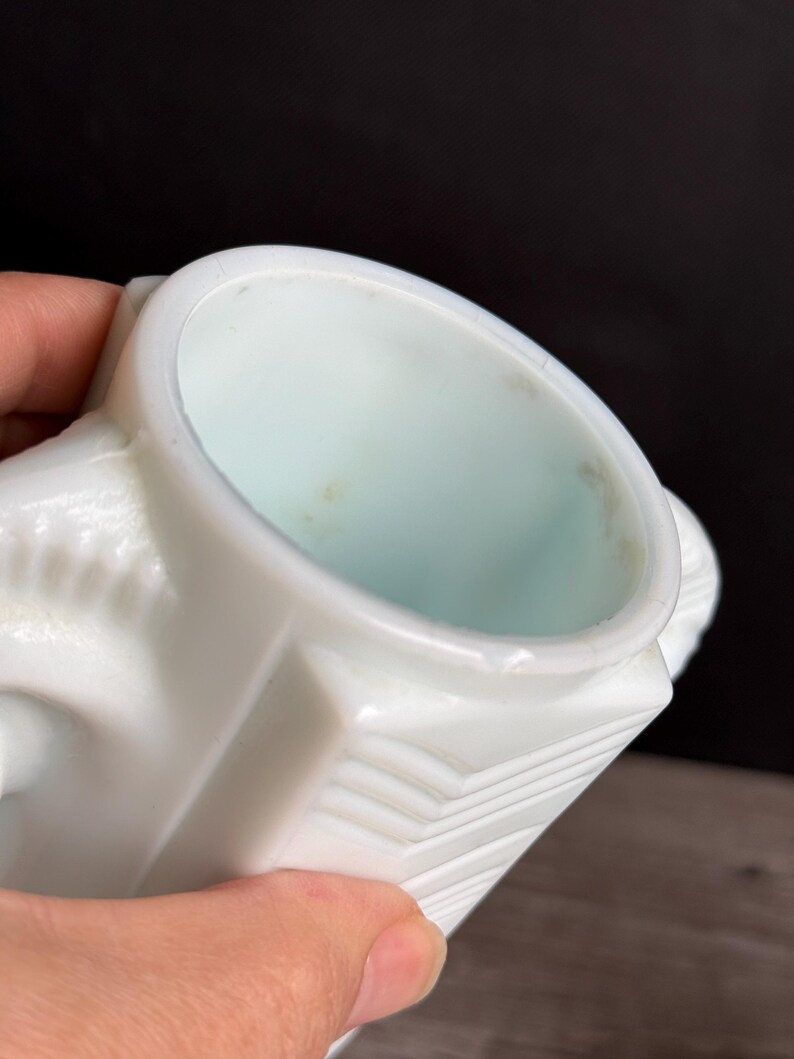 May include: A white, vintage-style creamer with a geometric design. The creamer has a square base and a rounded top, with a small handle on the side. The interior is a pale blue color.