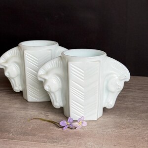 May include: Two white, art deco-style vases, each featuring three horse heads. The vases have a geometric design with a chevron pattern. A small sprig of purple flowers rests in front of the vases.