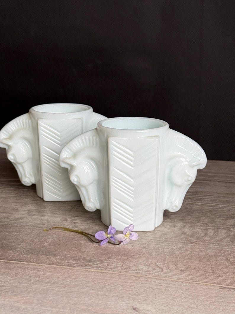 May include: Two white, art deco-style vases, each featuring three horse heads. The vases have a geometric design with a chevron pattern. A small sprig of purple flowers rests in front of the vases.