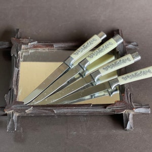 May include: Set of four silver knives with white handles featuring a floral design. The knives are displayed in a wooden frame with a mirror.