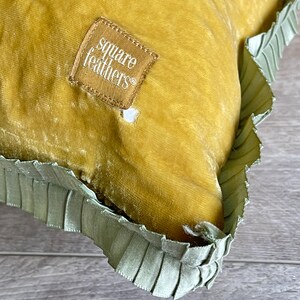 K&ouml;nnte beinhalten: Ein senfgelbes Samtkissen mit gr&uuml;nem R&uuml;schenbesatz. Das Kissen hat ein quadratisches Etikett mit der Aufschrift "square feathers".