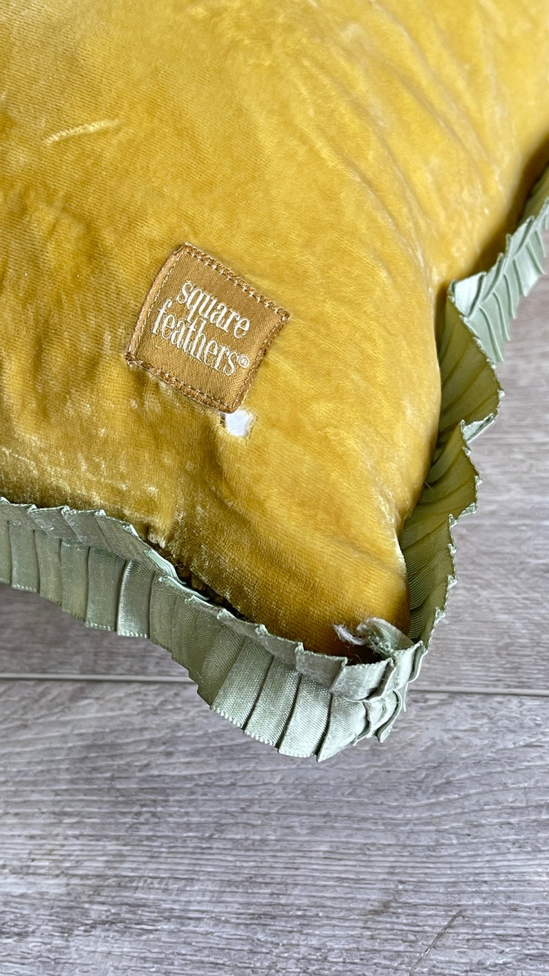K&ouml;nnte beinhalten: Ein senfgelbes Samtkissen mit gr&uuml;nem R&uuml;schenbesatz. Das Kissen hat ein quadratisches Etikett mit der Aufschrift "square feathers".