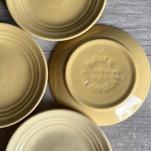 May include: A collection of yellow ceramic plates, some face up and some face down, with concentric ring detailing. The underside of one plate shows the text "BAUER MADE IN U.S.A. LOS ANGELES". The plates have a vintage aesthetic.