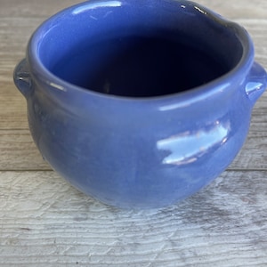 May include: A small, round, blue ceramic bowl with two small handles on either side. The bowl has a glossy finish and a smooth, curved shape. The color is a muted, periwinkle blue.